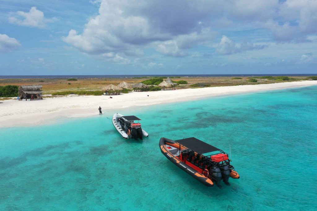 Two Powerboats float on clear turquoise water near Klein Curaçao’s sandy beach, dotted with thatched-roof huts and green brush. A person stands onshore under a blue, partly cloudy sky, as the ocean extends to the horizon.
