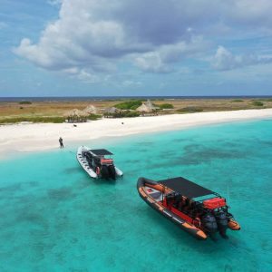 Twee motorboten van Klein Curaçao met de Powerboat dobberen op turquoise wateren nabij een wit zandstrand, met kleine rieten hutjes op de kust, een persoon in de buurt en lage begroeiing daarachter onder een gedeeltelijk bewolkte hemel.