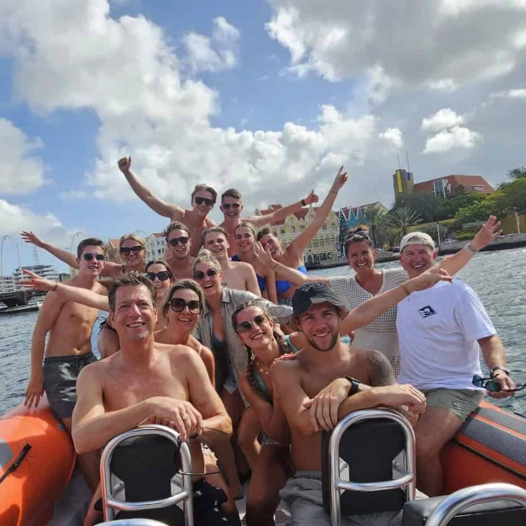 A group in swimwear smiles and poses on a Tugboat & Spanish Water orange inflatable boat. Most wear sunglasses, some raise their arms in excitement. Partly cloudy skies, buildings, and palm trees can be seen in the background.