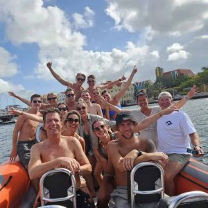 A group in swimwear smiles and poses on a Tugboat & Spanish Water orange inflatable boat. Most wear sunglasses, some raise their arms in excitement. Partly cloudy skies, buildings, and palm trees can be seen in the background.