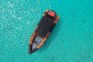 Aerial view of a Powerboat Private Charter with orange and black inflatable sides, covered seating, and a red container at the back floating on clear turquoise water with sunlight shimmering over the sea floor.
