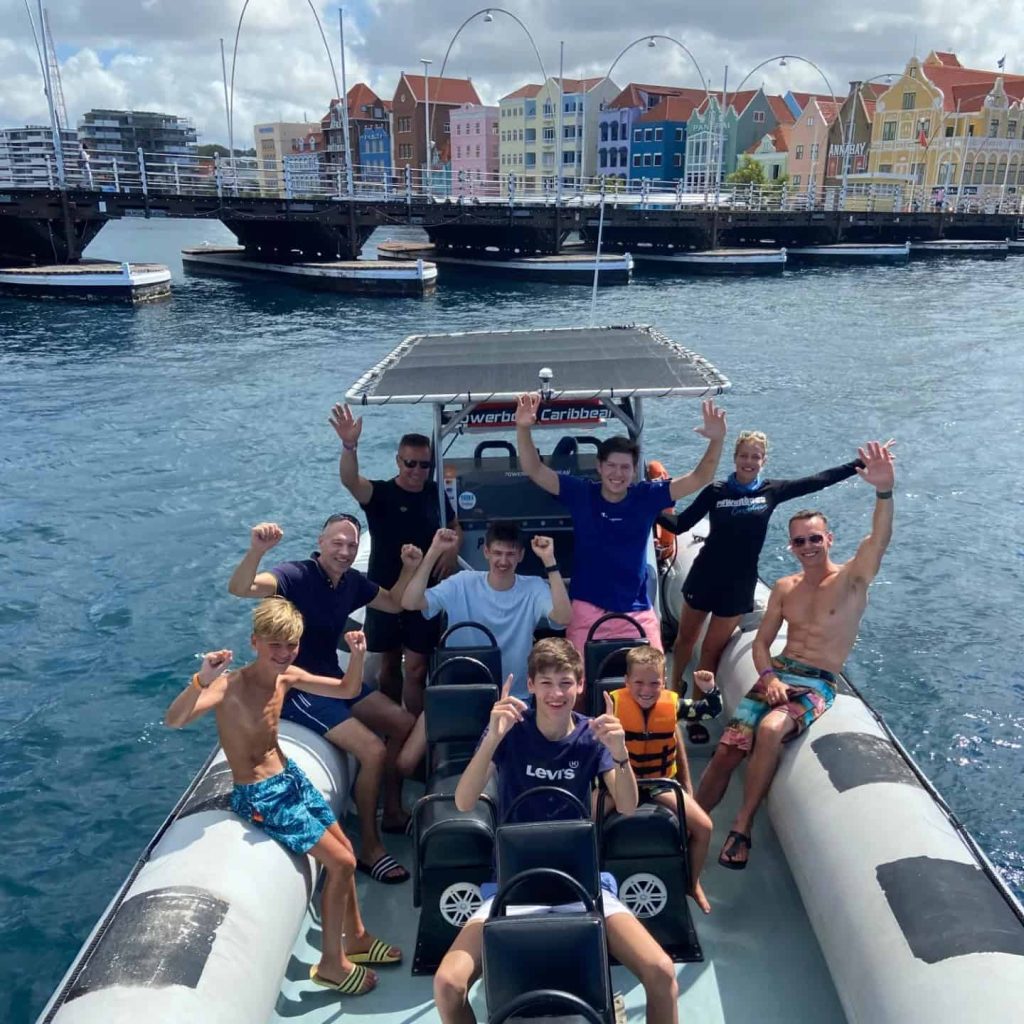 A group of smiling adults and children in swimwear enjoy the Westpunt – Sea Turtles & Blue Room tour, raising their arms excitedly on an inflatable boat with blue water, colorful waterfront buildings, and a bridge in the sunny background.