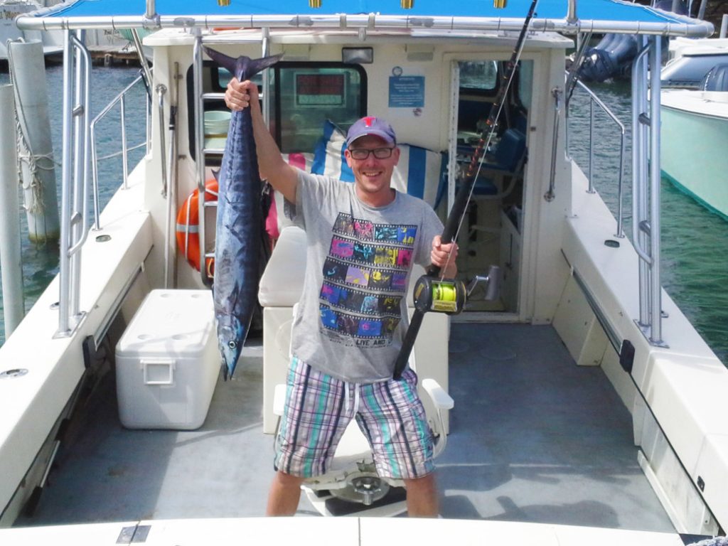 A smiling man in glasses stands on a docked boat, the Viking Sportsfisher 42ft, holding a fishing rod and a large fish. He wears casual gear, with water and other boats in the background.