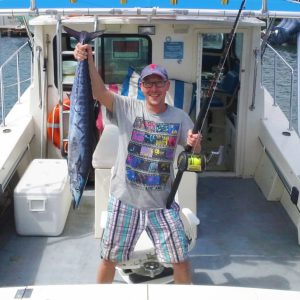 A smiling man in glasses stands on a docked boat, the Viking Sportsfisher 42ft, holding a fishing rod and a large fish. He wears casual gear, with water and other boats in the background.