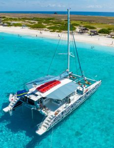 On the BlueFinn Charters Klein Curacao Trip, a white catamaran with patterned sides floats on turquoise water near a sandy beach. Guests relax onboard or swim nearby, with small huts, green shrubs, and ocean views under a blue sky.
