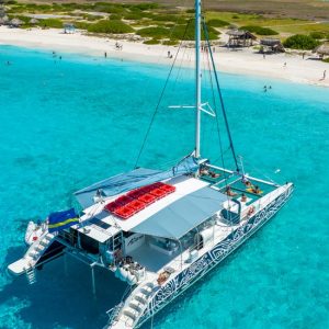 On the BlueFinn Charters Klein Curacao Trip, a white catamaran with patterned sides floats on turquoise water near a sandy beach. Guests relax onboard or swim nearby, with small huts, green shrubs, and ocean views under a blue sky.